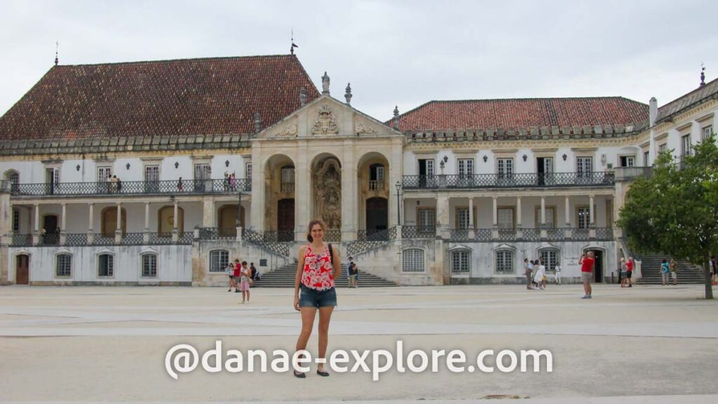 Eu no pátio da Universidade de Coimbra. Visto shorts jeans e blusa vermelha, ao fundo se veem os prédios da universidade