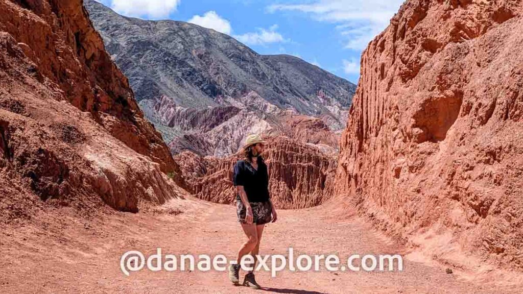 moça em shorts e blusa preta caminhando em meio a formações rochosas avermelhadas no Paseo de los Colorados, próximo ao Cerro de Las Siete Colores, em Purmamarca, norte da Argentina. Ao fundo, montanhas multicoloridas contrastam com o céu azul. 