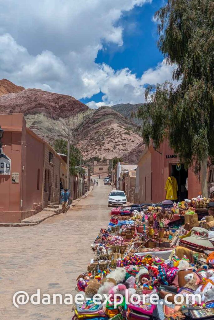 produtos artesanais andinos à venda em rua de Purmamarca, ao fundo casas em tons ocre e o Cerro de Los 7 Colores.