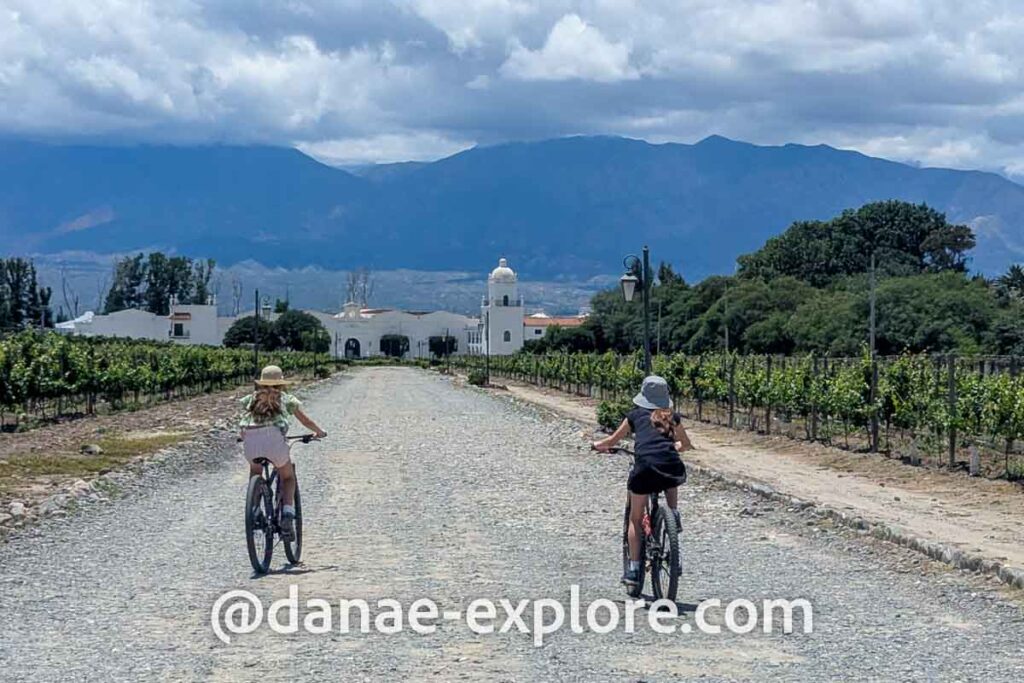 duas meninas andando de bicicleta em meio a vinhedos em Cafayate. Ao fundo, um casarão branco colonial sede de uma vinícola