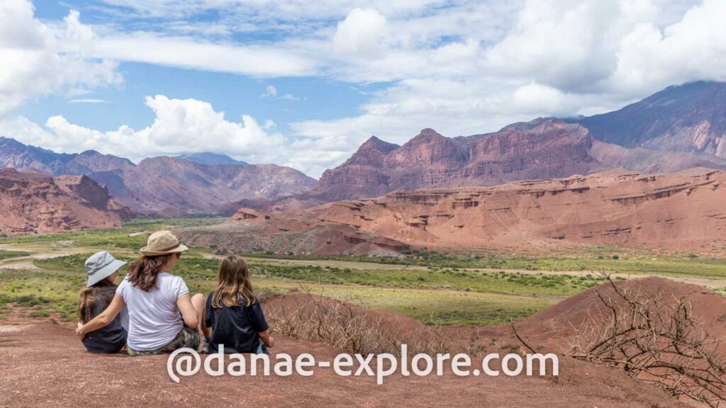 Mãe e duas filhas, sentadas de costas para câmera observam vale verde em meio a montanhas áridas, no Mirador 3 Cruces, a Ruta 68 entre Cafayate e Salta, parte de nosso roteiro pelo norte da Argentina