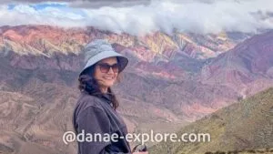 Vista das montanhas coloridas do Hornocal, com uma mulher sorridente em destaque, em cenário andino sob céu parcialmente nublado.
