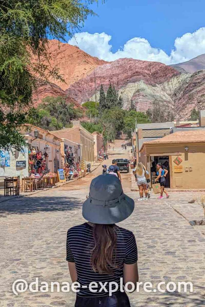 Menina de blusa preta e chapéu cinza, de costas para câmera, em rua de Purmarmarca, entre casas de adobe, lojas com produtos expostos e o Cerro de Los 7 Colores.