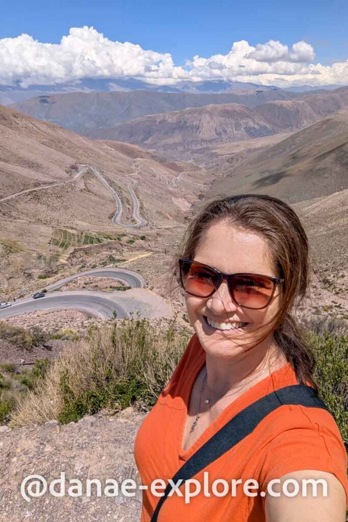 Selfie na Cuesta de Lipan, no caminho entre Purmamarca e Salinas Grandes, em dia de sol com algumas nuvens. A paisagem é desértica, com montanhas em ambos os lados e uma estrada sinuosa ao fundo