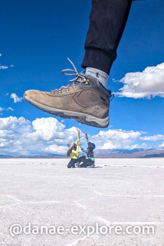 brincando com a perspectiva nas Salinas Grandes: foto mostra duas crianças sendo pisadas por um sapato gigante, em dia de sol com céu azul e nuvens