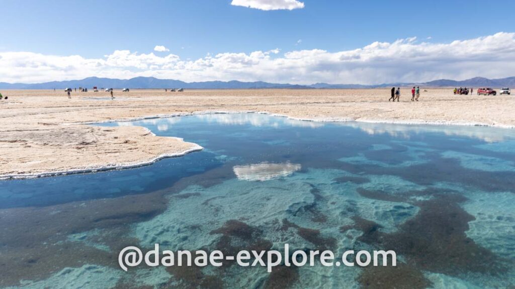 Lago de águas azuis em meio a Salinas Grandes. Há nuvens no céu, refletidas parcialmente no lago. Em segundo plano está o salar e algumas pessoas e veículos; ao fundo se veem as montanhas áridas características da região norte da Argentina.