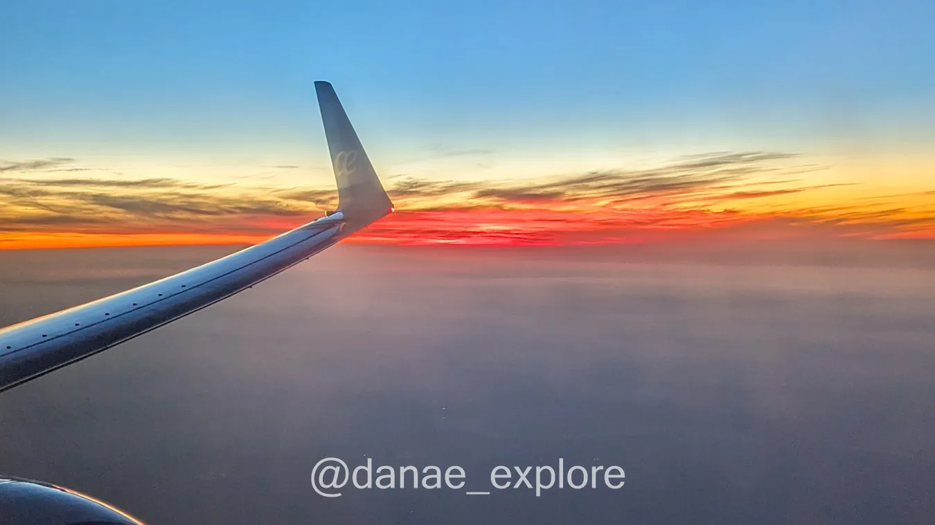Vista da janela de avião ao por-do-sol, é possível ver um pedaço da asa e o ceu em tons de azul e alaranjado, com nuvens abaixo