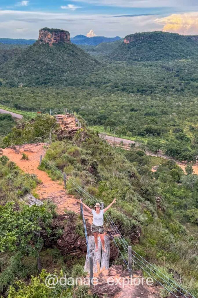 Moça em camiseta branca em trilha na Chapada das Mesas, em meio a vegetação baixa do cerrado, bastante verde. Ao fundo as formações rochosas características da Chapada das Mesas
