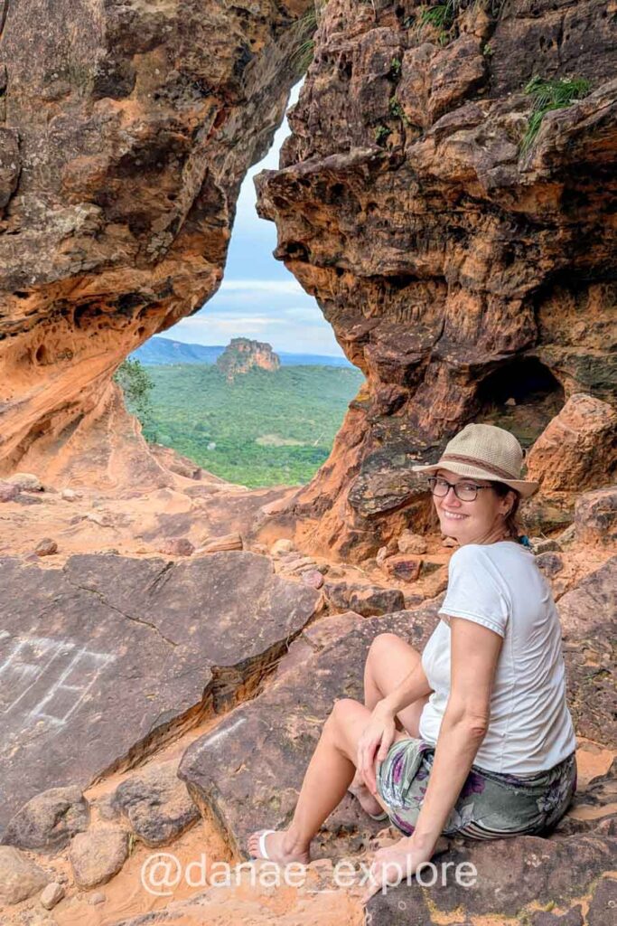 Moça de camiseta branca e chapéu olhando para câmera no Portal da Chapada das Mesas, por uma fresta na parede rochosa se vê uma das formações características da Chapada