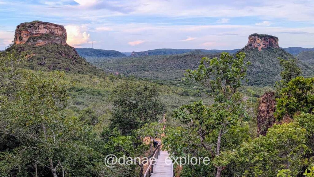 Vista de mirante na Chapada das Mesas. Há duas formações rochosas em cada lado da imagem e uma passarela em madeira conduzindo ao caminho por entre elas. A vegeração verde do cerrado cobre toda a cena.