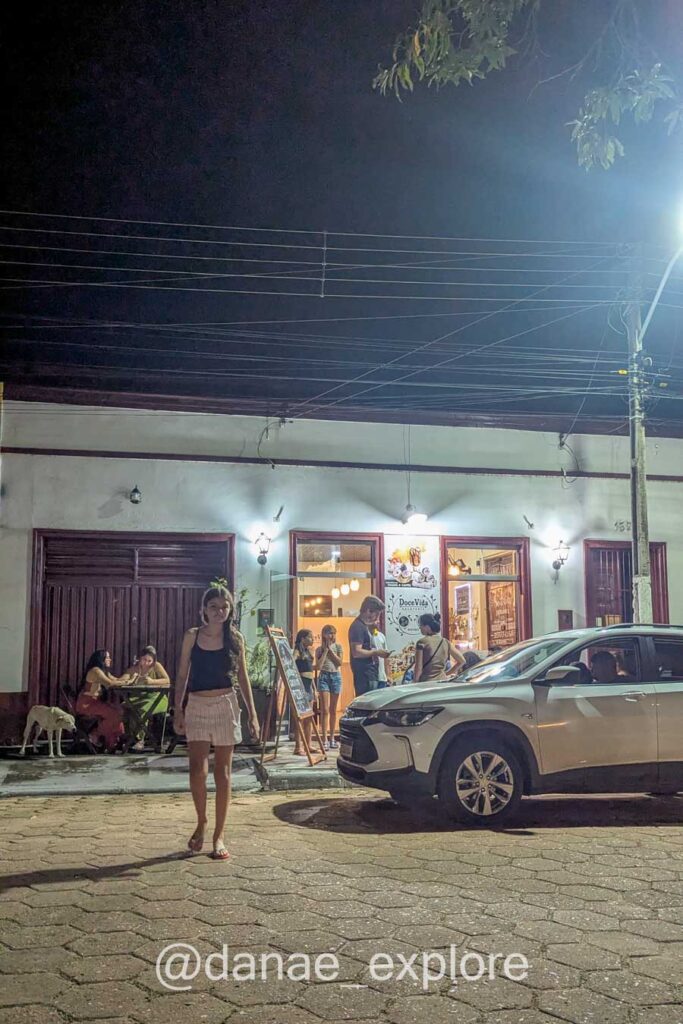 Menina caminha em direção à câmera, ao fundo sorveteria na cidade de Carolina. É uma noite de calor. Há um carro estacionado parcialmente visível, e pessoas ao fundo na calçada