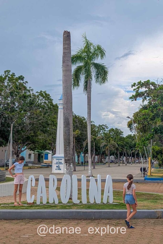 Duas adolescentes caminham perto de letreiro onde se lê CAROLINA, em praça na cidade de Carolina no Maranhão. É um dia quente e nublado.