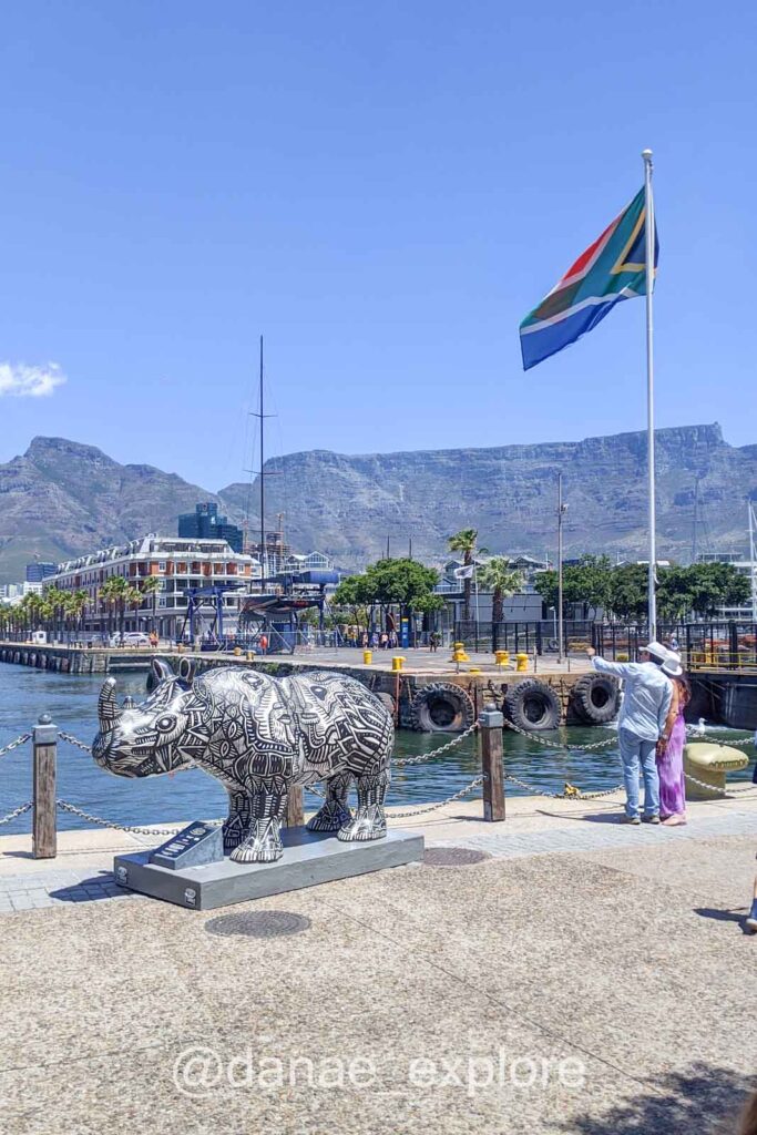 Escultura de rinoceronte no Victoria and Alfred Waterfront. Há também um casal ao lado de um mastro com a bandeira da África do Sul.