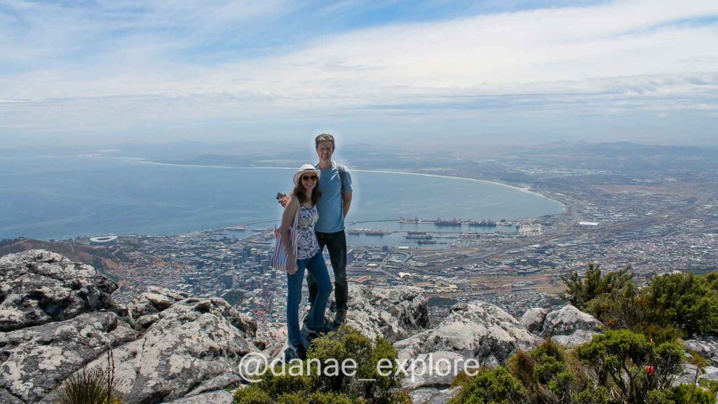 Casal no topo da Table Mountain, abaixo ao fundo se vê a Cidade do Cabo em dia de céu azul com muitas nuvens e alguma névoa