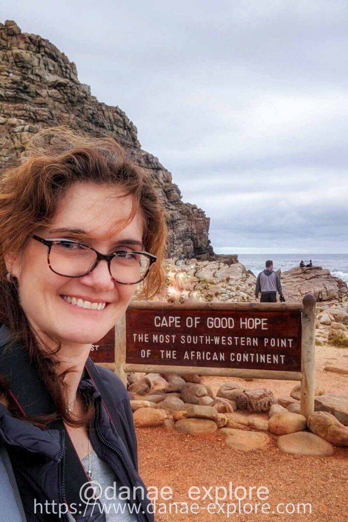 selfie de moça branca de óculos e cabelos ao vento em frente a placa do Cabo da Boa Esperança, em dia nublado