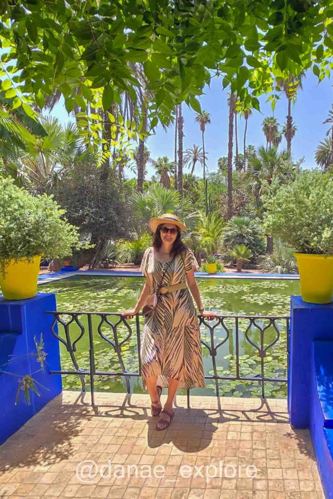 Eu, usando vestido e chapéu, sorrio para câmera em frente a espelho d'água no Jardim Majorelle, em Marrrakesh