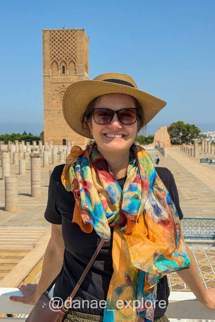 Me smiling at the camera, wearing a colorful scarf, sunglasses, and a hat, with the Hassan Tower in Rabat in the background