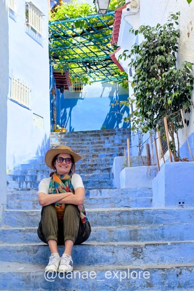 lenço e chapéu, olha para câmera sorrindo em ruela com casas azuis, na cidade azul do Marrocos, Chefchaouen, partte de nosso roteiro de 10 dias no Marrocoslenço e chapéu, olha para câmera sorrindo em escadaria rodeada por casas azuis, na cidade azul do Marrocos, Chefchaouen, partte de nosso roteiro de 10 dias no Marrocos