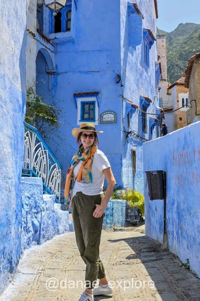 Mulher em calça e camiseta, com lenço e chapéu, olha para câmera sorrindo em ruela com casas azuis, na cidade azul do Marrocos, Chefchaouen - sugestão de o que vestir no Marrocos