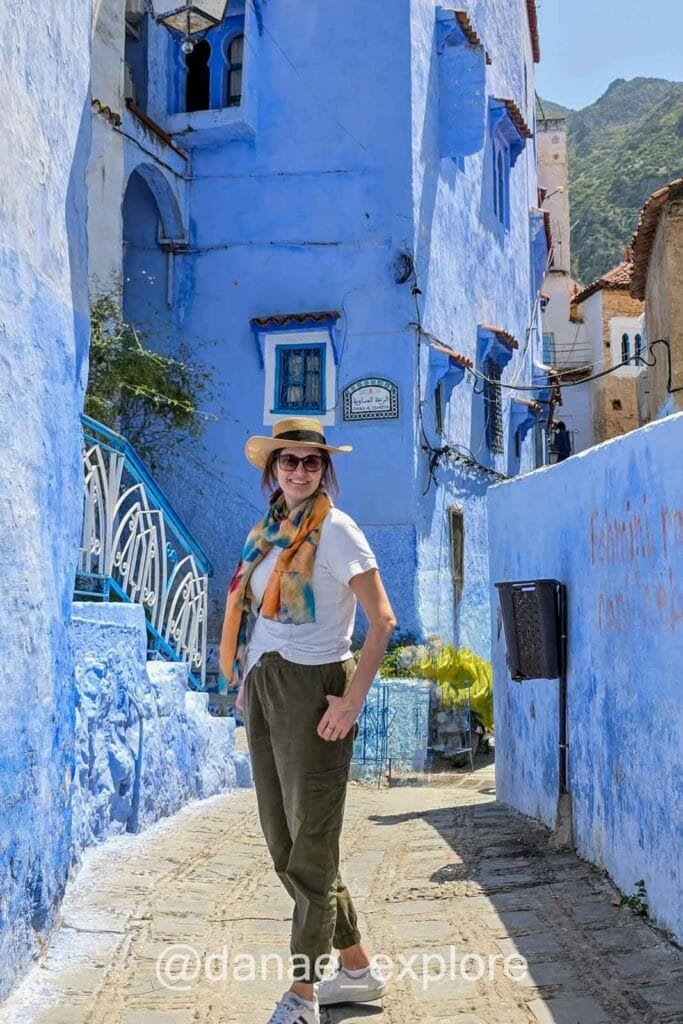 Woman wearing pants and a T-shirt, with a scarf and hat, smiling at the camera in a narrow alley lined with blue houses in Chefchaouen, Morocco—the Blue City; outfit idea for what to wear in Morocco