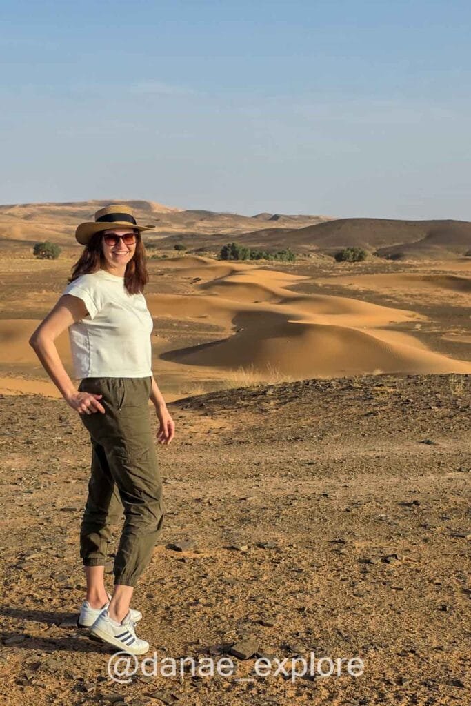 Me, wearing long pants, a T-shirt, and a hat, standing on top of a sand dune in the Sahara Desert near Merzouga on a clear blue-sky day