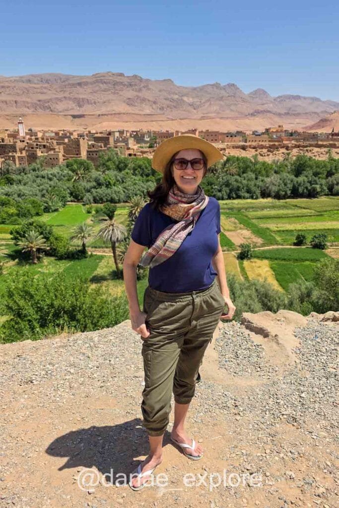 Woman in pants and a T-shirt smiling at the camera at a viewpoint, overlooking a lush green valley with a kasbah of adobe buildings, with the mountains of the Moroccan desert in the background