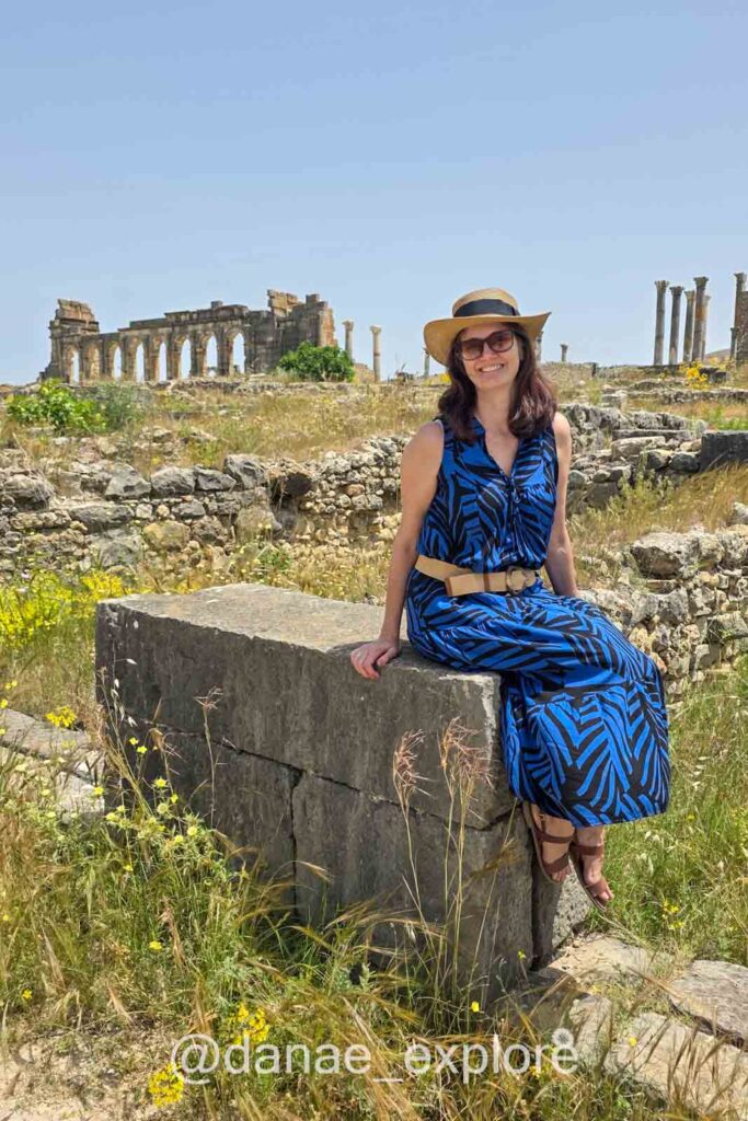 Moça em vestido longo azul estampado, óculos de sol e chapéu sentada em banco de pedra nas ruínas de Volubilis - dicas do que vestir no Marrocos