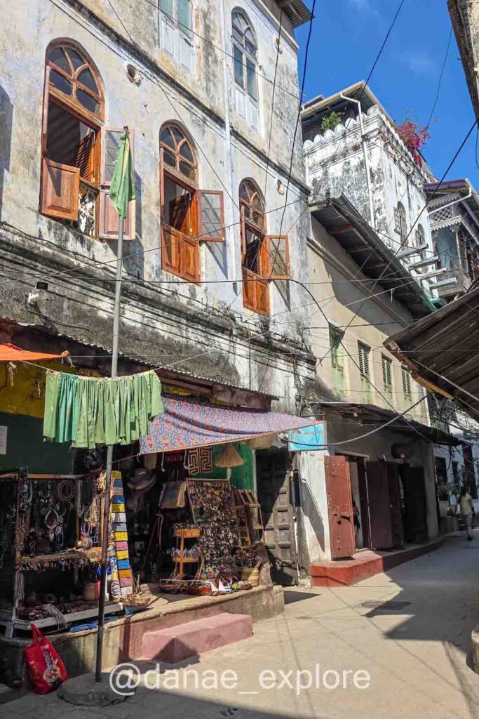 ruela em Stone Town com prédios antigos e lojas de souvenirs no térreo. Há sinais de desgaste nas edificações