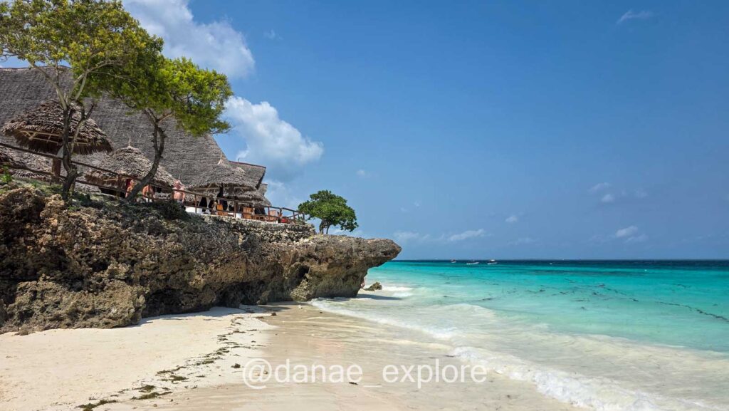 Nungwi Beach em Zanzibar em dia de céu azul. Há um rochedo com uma construção em teto de palha a esquerda e algumas árvores, e o mar em tom azul turquesa a direita