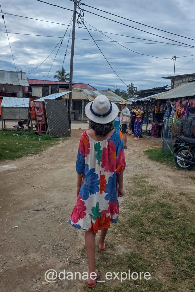 moça em vestido estampado colorido e chapéu branco caminha por rua de terra na vila de Nungwi, em Zanzibar