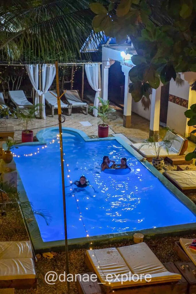 Foto noturna da piscina do hotel Kilimanjaro, onde nos hospedamos em  Nungwi, Zanzibar. A piscina está iluminada, há algumas cadeiras ao redor e três pessoas nadando 