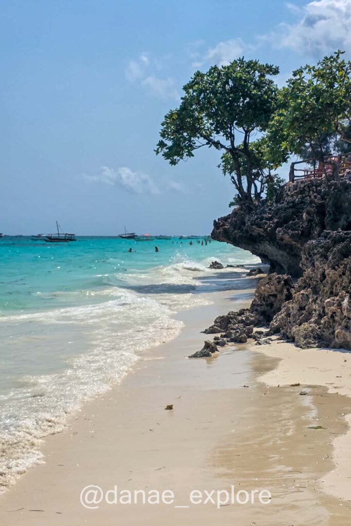 praia em Nungwi, Zanzibar. O mar é azul turquesa, há alguns barcos pequenos ao fundo e pessoas nadando, em primeiro plano se vê a areia branca, há pedras com árvores no canto direito.