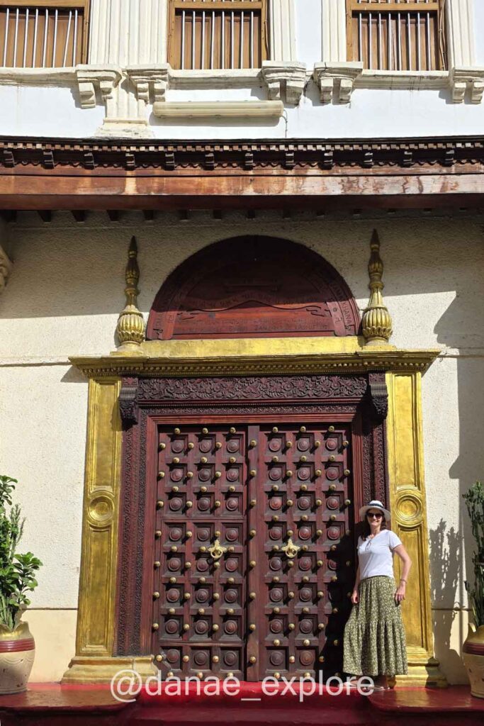 Mulher em saia longa, camiseta e chapéu em frente a porta decorada em Stone Town, parte de nossas sugestões do que fazer em Zanzibar