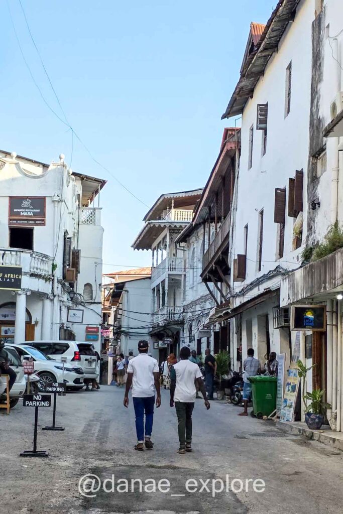 pessoas caminham em rua de Stone Town, em meio a lojas e casas de três pavimentos brancas, algumas com sacadas de madeira