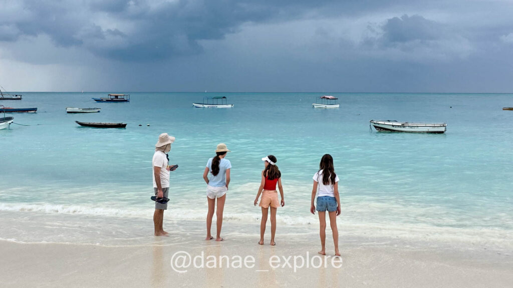 quatro olhando o mar azul em Zanzibar, em dia nublado. Há diversos pequenos barcos na água. A areia é branca bem clara e as ondas suave