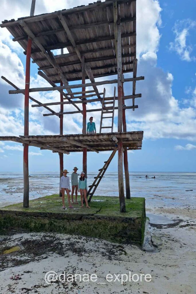 Plataforma de madeira com três pavimentos na praia de Bwejuu - Paje durante a maré baixa, não é possível avistar o oceano, apenas a areia com algumas pequenas poças de água. Há três pessoas no primeiro patamar da plataforma e uuma no segundo