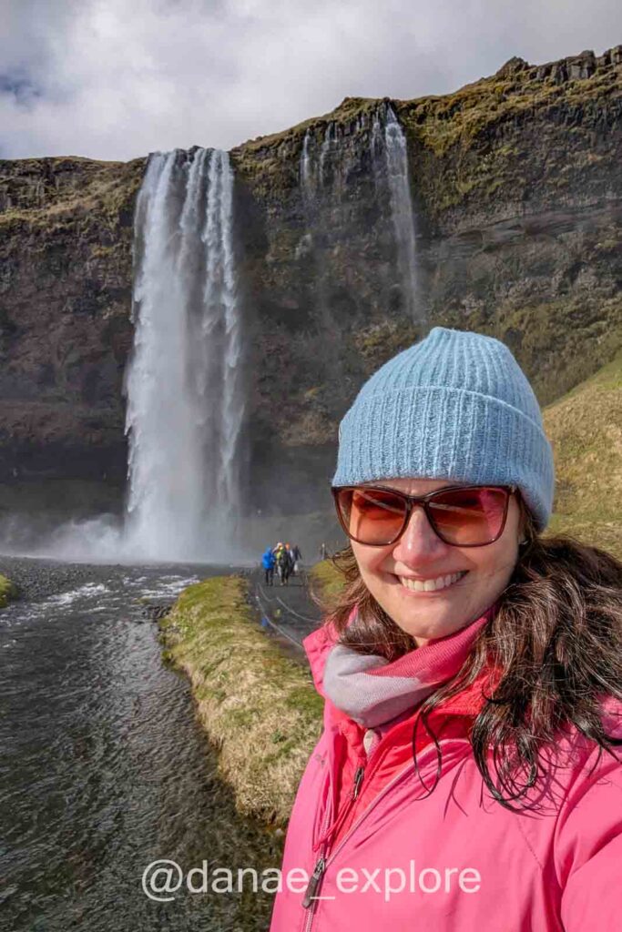 Selfie sorrindo, com gorro azul e jaqueta rosa, em frente à cachoeira Seljalandsfoss, na costa sul da Islândia. A água despenca de um alto penhasco coberto de musgo, em um dia com muitas nuvens no céu