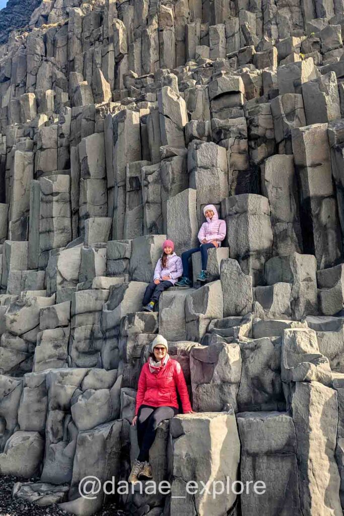 Três pessoas em roupas de frio, com casaco e gorro, sentadas entre as formações hexagonais de basalto na praia de Reynisfjara, na Islândia