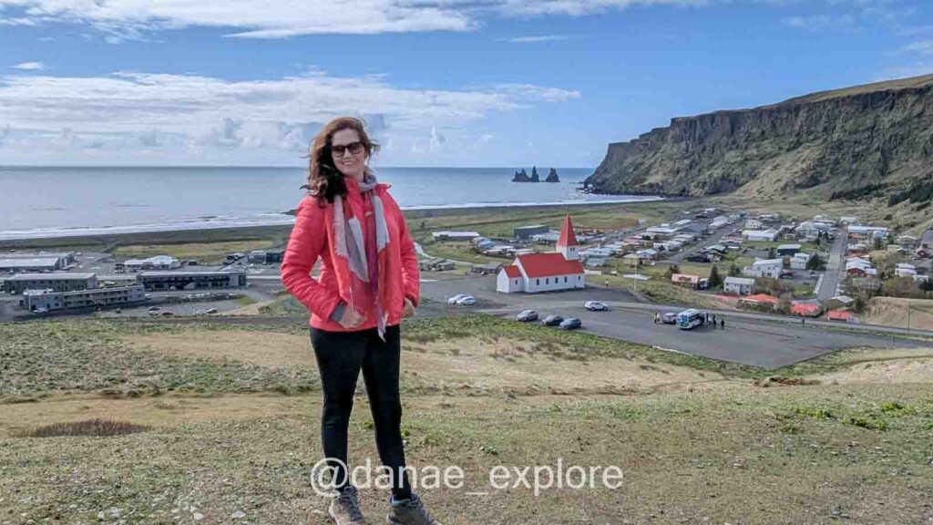 Uma mulher com jaqueta vermelha e cachecol posa sorrindo em um mirante com vista para a vila de Vík í Mýrdal, na Islândia. Ao fundo, destacam-se a igreja de telhado vermelho, o vilarejo costeiro e as formações rochosas Reynisdrangar no mar.