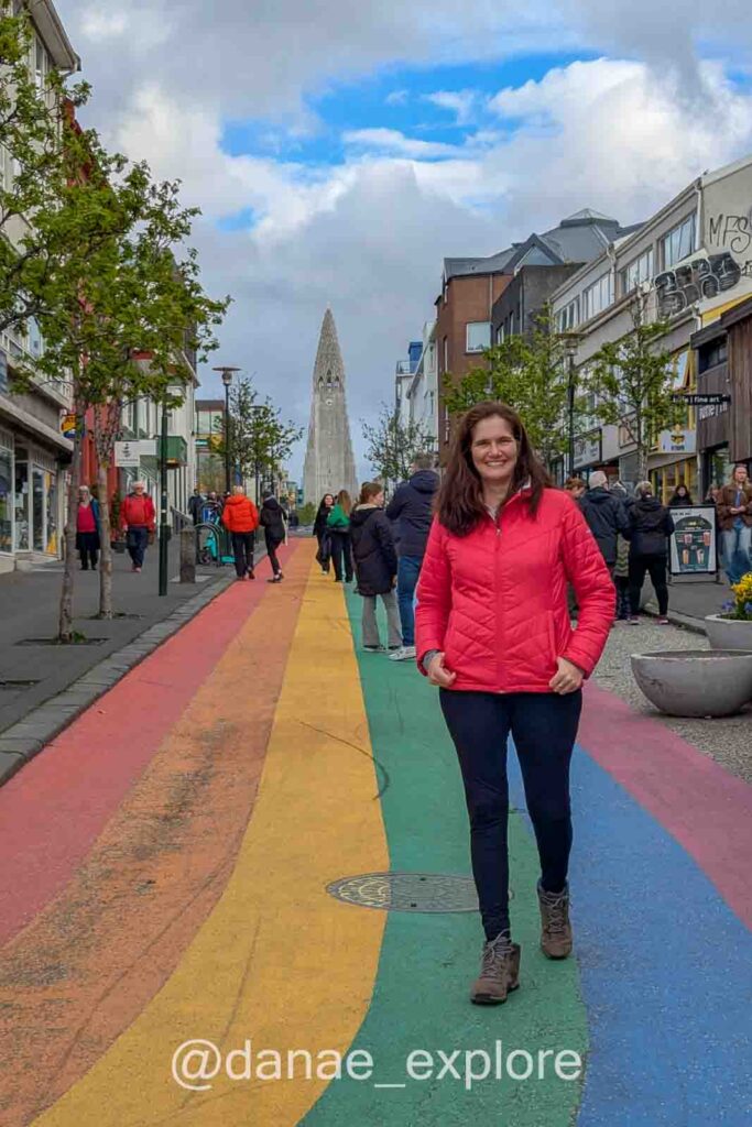 Eu, com jaqueta vermelha, caminhando por uma rua com as cores do arco-íris pintada no chão, no centro de Reykjavík, primeira parada de nosso roteiro pela Islândia. Ao fundo, destaca-se a icônica igreja Hallgrímskirkja