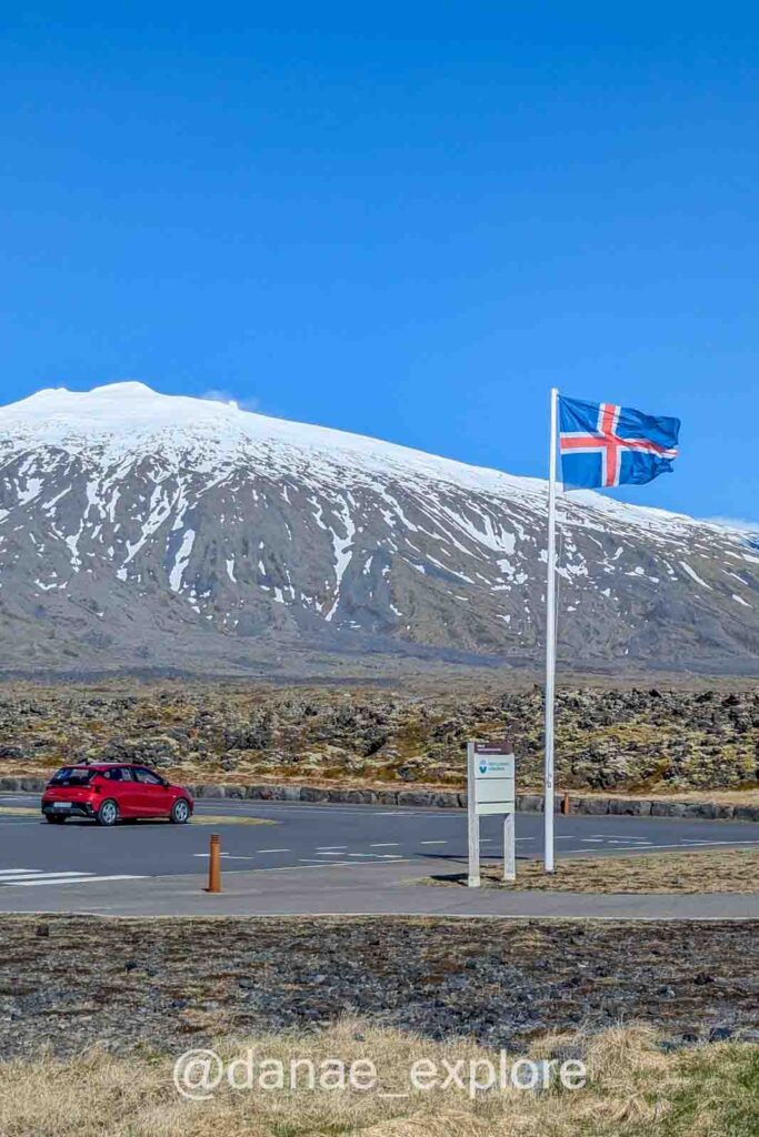 Bandeira da Islândia tremula ao vento diante do vulcão Snæfellsjökull, na Islândia, cujo topo está coberto de neve. Um carro vermelho cruza a estrada sob o céu azul.