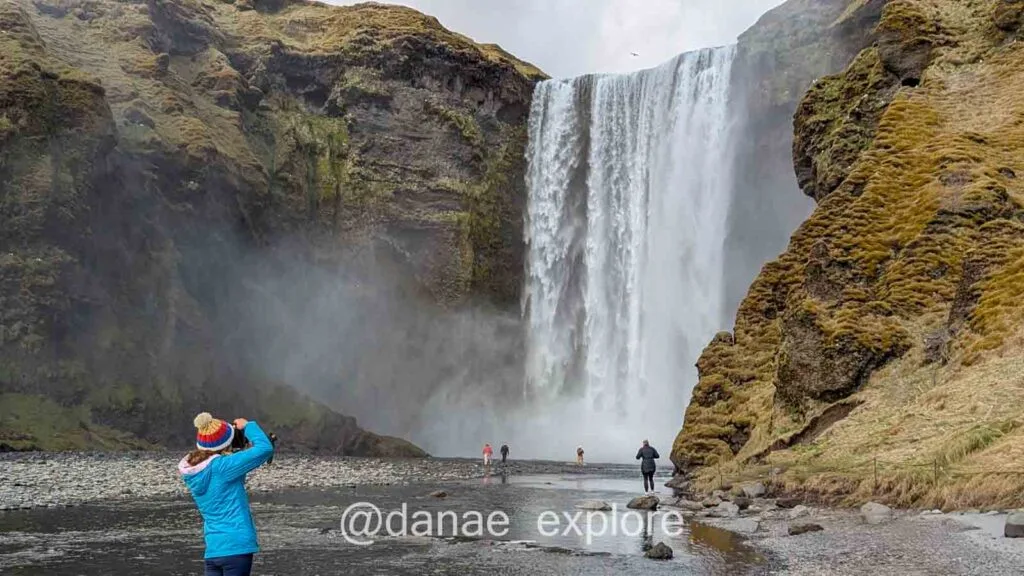 A person in a blue jacket and colorful hat photographs Skógafoss waterfall, one of the must-see attractions on an Iceland itinerary. The waterfall tumbles between moss-covered cliffs, creating a mist that envelops tourists walking along the rocky shore.