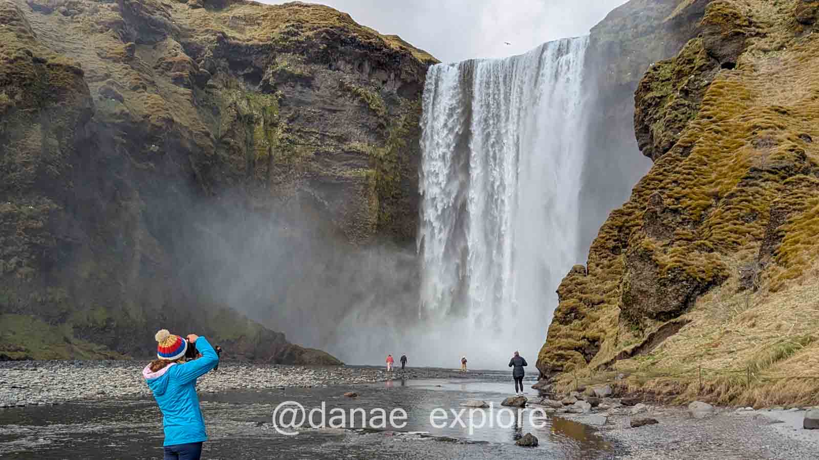 Uma pessoa com jaqueta azul e gorro colorido fotografa a cachoeira Skógafoss, uma das atrações imperdíveis em um roteiro pela Islândia. A queda d’água despenca entre penhascos cobertos de musgo, criando uma névoa que envolve turistas caminhando pela margem rochosa