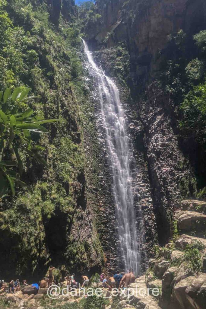Cachoeira do Segredo, próxima a São Jorge, em dia de sol. Há diversos visitantes nas pedras na base da cachoeira