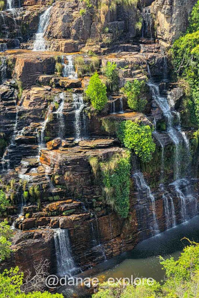 Vista da Cachoeira Almécegas I, uma imponente cachoeira com múltiplas quedas descendo entre rochas alaranjadas e vegetação verde, formando poços ao longo da parede rochosa na Chapada dos Veadeiros.