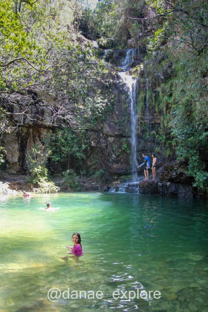 Crianças e adultos nadam em um poço de água verde esmeralda ao pé de uma cachoeira fina, cercada por mata na Chapada dos Veadeiros.