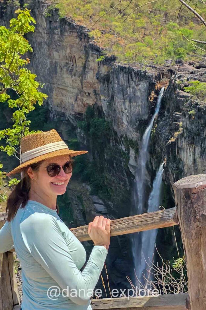 Eu, com chapéu e óculos escuros, olho para câmera; ao fundo duas cachoeiras altas, os Saltos do Rio Preto, na trilha amarela do Parque Nacional da Chapada dos Veadeiros