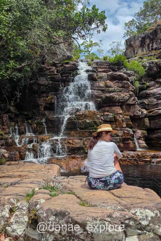 Eu sentada numa pedra em frente a Cachoeira Arcanjos. Estou usando shorts, camiseta e chapéu, é um dia com sol e muitas nuvens