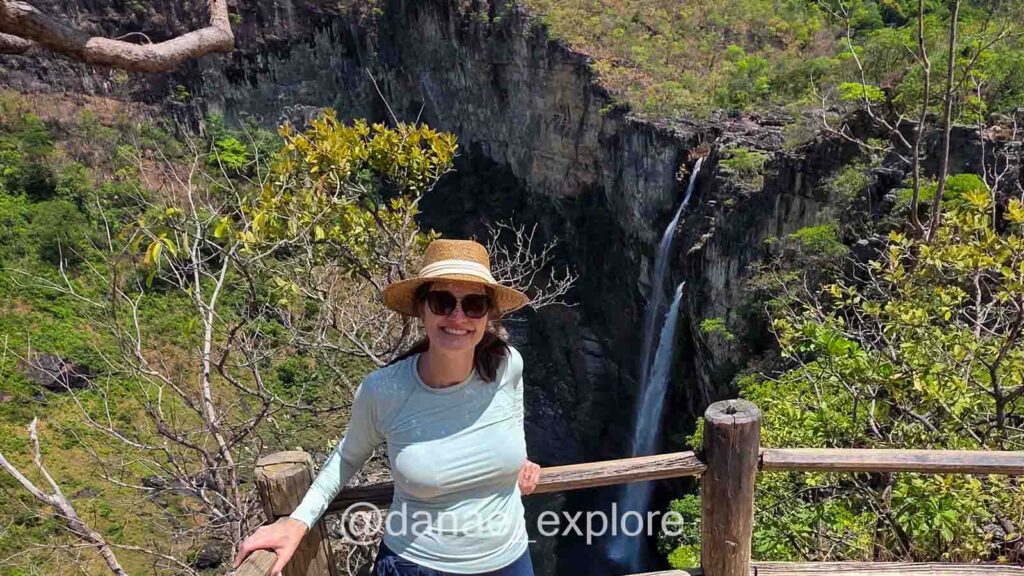 Mulher sorridente com chapéu e óculos escuros posa em mirante com vista para uma cachoeira alta e estreita (Saltos do Rio Preto) cercada por vegetação na Chapada dos Veadeiros.