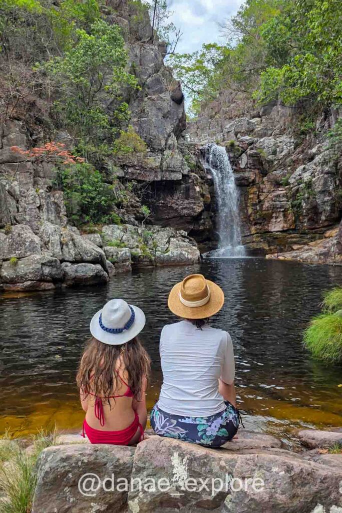 Eu e minha filha, ambas de chapéu, sentadas de costas, observamos a Cachoeira dos Anjos, que deságua em um poço escuro rodeado por rochas e vegetação nativa, na Chapada dos Veadeiros